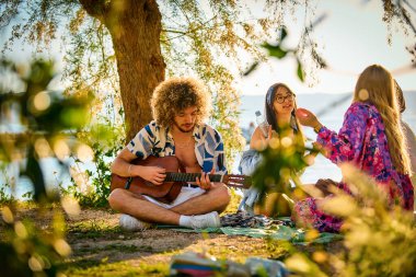 Bir grup arkadaş plajda toplanıp müzik, kahkaha ve birlikteliğin sıcaklığıyla dolu neşeli anları paylaşıyorlar. Gençler sahilde eğleniyor.