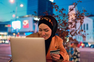 A woman wearing a hijab sitting in a cozy coffee at night, shopping online on her laptop and making a purchase with her credit card, surrounded by a calm, modern atmosphere clipart