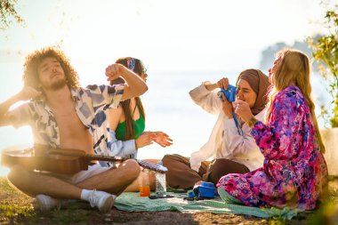 Bir grup arkadaş plajda toplanıp müzik, kahkaha ve birlikteliğin sıcaklığıyla dolu neşeli anları paylaşıyorlar. Gençler sahilde eğleniyor.
