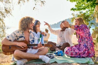 Bir grup arkadaş plajda toplanıp müzik, kahkaha ve birlikteliğin sıcaklığıyla dolu neşeli anları paylaşıyorlar. Gençler sahilde eğleniyor.