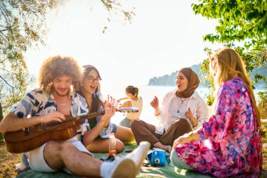 Bir grup arkadaş plajda toplanıp müzik, kahkaha ve birlikteliğin sıcaklığıyla dolu neşeli anları paylaşıyorlar. Gençler sahilde eğleniyor.