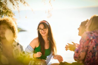 Bir grup arkadaş plajda toplanıp müzik, kahkaha ve birlikteliğin sıcaklığıyla dolu neşeli anları paylaşıyorlar. Gençler sahilde eğleniyor.