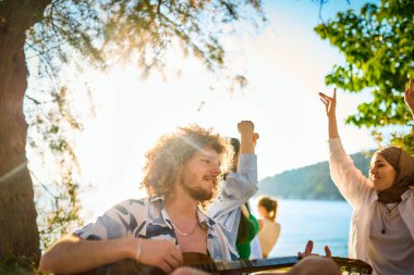 Bir grup arkadaş plajda toplanıp müzik, kahkaha ve birlikteliğin sıcaklığıyla dolu neşeli anları paylaşıyorlar. Gençler sahilde eğleniyor.