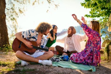 Bir grup arkadaş plajda toplanıp müzik, kahkaha ve birlikteliğin sıcaklığıyla dolu neşeli anları paylaşıyorlar. Gençler sahilde eğleniyor.