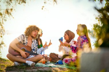 Bir grup arkadaş plajda toplanıp müzik, kahkaha ve birlikteliğin sıcaklığıyla dolu neşeli anları paylaşıyorlar. Gençler sahilde eğleniyor.