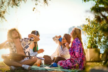 Bir grup arkadaş plajda toplanıp müzik, kahkaha ve birlikteliğin sıcaklığıyla dolu neşeli anları paylaşıyorlar. Gençler sahilde eğleniyor.