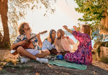 Bir grup arkadaş plajda toplanıp müzik, kahkaha ve birlikteliğin sıcaklığıyla dolu neşeli anları paylaşıyorlar. Gençler sahilde eğleniyor.