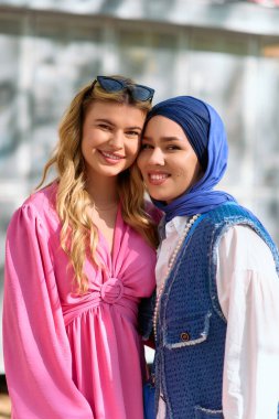 İki arkadaş, biri tesettür içinde, diğeri ise çarpıcı bir sarışın, güneşin altında neşeli bir anı paylaşıyorlar, çeşitliliğin ve arkadaşlığın güzelliğini somutlaştırıyorlar..