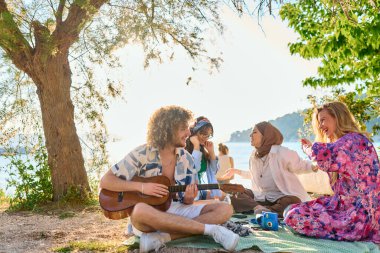 Bir grup arkadaş plajda toplanıp müzik, kahkaha ve birlikteliğin sıcaklığıyla dolu neşeli anları paylaşıyorlar. Gençler sahilde eğleniyor.