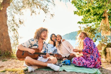Bir grup arkadaş plajda toplanıp müzik, kahkaha ve birlikteliğin sıcaklığıyla dolu neşeli anları paylaşıyorlar. Gençler sahilde eğleniyor.