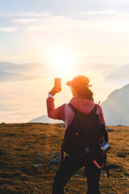 Bir kadın yürüyüşçü, sosyal medya için dağın zirvesinden gün batımını yakalar. Bir kadın yürüyüşçü, dağ zirvesinde günbatımının keyfini çıkararak sosyal medya için akıllı telefonuyla o anı yakalar.