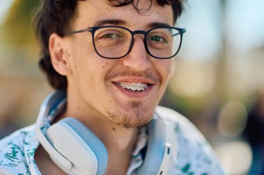 Gözlük ve kulaklık takan neşeli genç bir adam sıcak, güneşli bir açık havada ışıl ışıl gülümsüyor, kendine güven ve pozitiflik saçıyor.. 