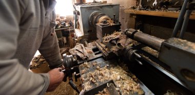 Aletler, ahşap kalıplar ve endüstriyel ekipmanlarla dolu kırsal ve dağınık bir atölyede makinelerle çalışan yaşlı bir zanaatkar. Sıcak güneş ışığı sahneye nostaljik ve klasik bir his katıyor.