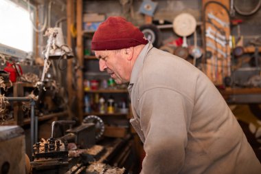 Aletler, ahşap kalıplar ve endüstriyel ekipmanlarla dolu kırsal ve dağınık bir atölyede makinelerle çalışan yaşlı bir zanaatkar. Sıcak güneş ışığı sahneye nostaljik ve klasik bir his katıyor.