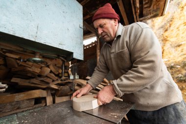 Bir marangoz elinin, kaba ahşap bir kalası, kırsal bir atölyedeki testerenin içinden geçirdiği yakın çekim. Görüntü, marangozluk, hassasiyet ve ahşap işçiliğindeki sıkı çalışmayı yakalıyor.