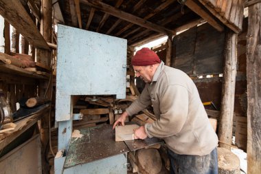 Bir marangoz elinin, kaba ahşap bir kalası, kırsal bir atölyedeki testerenin içinden geçirdiği yakın çekim. Görüntü, marangozluk, hassasiyet ve ahşap işçiliğindeki sıkı çalışmayı yakalıyor.