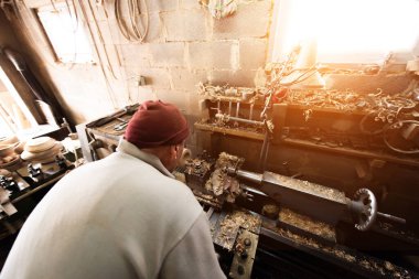 Aletler, ahşap kalıplar ve endüstriyel ekipmanlarla dolu kırsal ve dağınık bir atölyede makinelerle çalışan yaşlı bir zanaatkar. Sıcak güneş ışığı sahneye nostaljik ve klasik bir his katıyor.