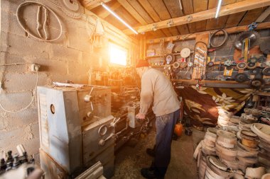 Aletler, ahşap kalıplar ve endüstriyel ekipmanlarla dolu kırsal ve dağınık bir atölyede makinelerle çalışan yaşlı bir zanaatkar. Sıcak güneş ışığı sahneye nostaljik ve klasik bir his katıyor.