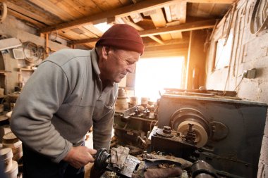 Aletler, ahşap kalıplar ve endüstriyel ekipmanlarla dolu kırsal ve dağınık bir atölyede makinelerle çalışan yaşlı bir zanaatkar. Sıcak güneş ışığı sahneye nostaljik ve klasik bir his katıyor.