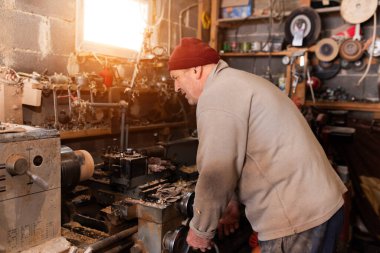 Aletler, ahşap kalıplar ve endüstriyel ekipmanlarla dolu kırsal ve dağınık bir atölyede makinelerle çalışan yaşlı bir zanaatkar. Sıcak güneş ışığı sahneye nostaljik ve klasik bir his katıyor.