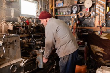 Aletler, ahşap kalıplar ve endüstriyel ekipmanlarla dolu kırsal ve dağınık bir atölyede makinelerle çalışan yaşlı bir zanaatkar. Sıcak güneş ışığı sahneye nostaljik ve klasik bir his katıyor.