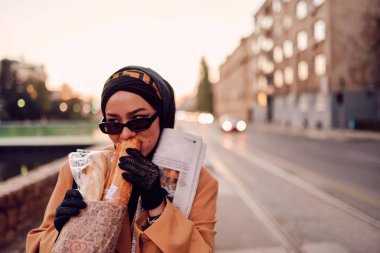 Güzel bir Arap kadını, modern bir tesettüre bürünmüş, kentin sokaklarında güvenle yürüyor, bir çanta ve gazete taşıyor.