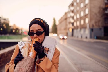 Güzel bir Arap kadını, modern bir tesettüre bürünmüş, kentin sokaklarında güvenle yürüyor, bir çanta ve gazete taşıyor.