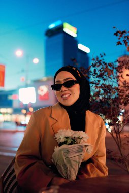 Şık bir tesettür ve güneş gözlüğü takmış güzel bir Arap kadını geceleri modern bir şehir kafesinin kalbinde güven içinde oturuyor.