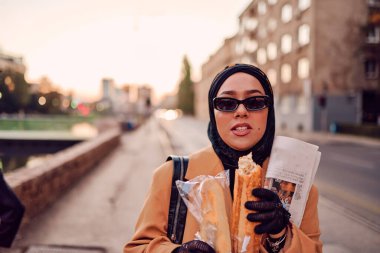 Güzel bir Arap kadını, modern bir tesettüre bürünmüş, kentin sokaklarında güvenle yürüyor, bir çanta ve gazete taşıyor.