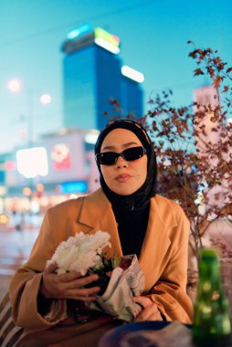 Şık bir tesettür ve güneş gözlüğü takmış güzel bir Arap kadını geceleri modern bir şehir kafesinin kalbinde güven içinde oturuyor.