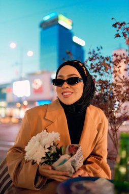 Şık bir tesettür ve güneş gözlüğü takmış güzel bir Arap kadını geceleri modern bir şehir kafesinin kalbinde güven içinde oturuyor.