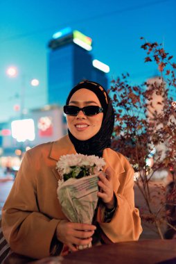 Şık bir tesettür ve güneş gözlüğü takmış güzel bir Arap kadını geceleri modern bir şehir kafesinin kalbinde güven içinde oturuyor.