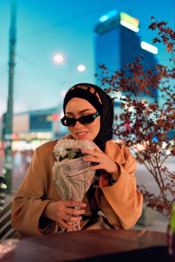 Şık bir tesettür ve güneş gözlüğü takmış güzel bir Arap kadını geceleri modern bir şehir kafesinin kalbinde güven içinde oturuyor.