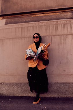 Kentte otobüs bekleyen, elinde bir somun ekmek tutarken gazete okuyan, tesettürlü modern bir Arap kadın..