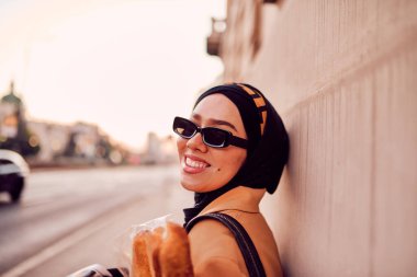 Kentte otobüs bekleyen, elinde bir somun ekmek tutarken gazete okuyan, tesettürlü modern bir Arap kadın..