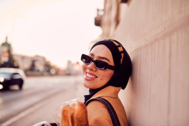 Kentte otobüs bekleyen, elinde bir somun ekmek tutarken gazete okuyan, tesettürlü modern bir Arap kadın..