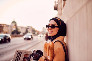 Kentte otobüs bekleyen, elinde bir somun ekmek tutarken gazete okuyan, tesettürlü modern bir Arap kadın..