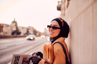 Kentte otobüs bekleyen, elinde bir somun ekmek tutarken gazete okuyan, tesettürlü modern bir Arap kadın..