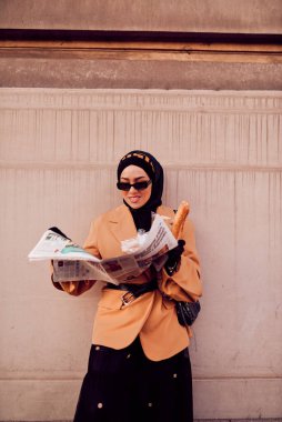 Kentte otobüs bekleyen, elinde bir somun ekmek tutarken gazete okuyan, tesettürlü modern bir Arap kadın..