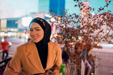 Güzel, gülümseyen bir Arap kadın gece vakti şehirde bir kahvede oturuyor, şehir ortamının coşkulu atmosferinden zevk alırken zarafet ve neşe saçıyor.