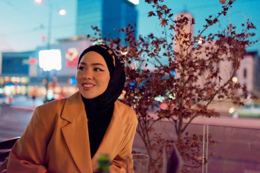 Güzel, gülümseyen bir Arap kadın gece vakti şehirde bir kahvede oturuyor, şehir ortamının coşkulu atmosferinden zevk alırken zarafet ve neşe saçıyor.