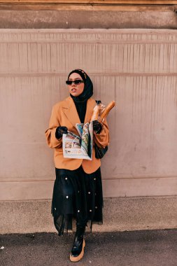 Kentte otobüs bekleyen, elinde bir somun ekmek tutarken gazete okuyan, tesettürlü modern bir Arap kadın..