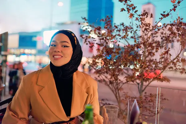 Güzel, gülümseyen bir Arap kadın gece vakti şehirde bir kahvede oturuyor, şehir ortamının coşkulu atmosferinden zevk alırken zarafet ve neşe saçıyor.