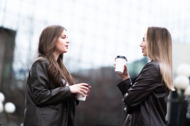 Deri ceket giymiş iki genç kadın dışarıda kahkaha atıyor ve kahveden zevk alıyorlar. Arka planda modern cam binalar olan dostane şehir manzarası.