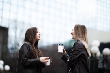 Deri ceket giymiş iki genç kadın dışarıda kahkaha atıyor ve kahveden zevk alıyorlar. Arka planda modern cam binalar olan dostane şehir manzarası.