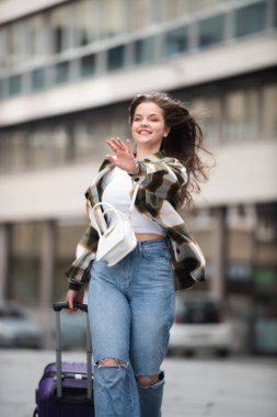Yırtık kotlu mutlu genç turist kadın kaldırımda bavuluyla yürüyor. Genç Kadın Bavulla Neşeli Yürüyüş. 