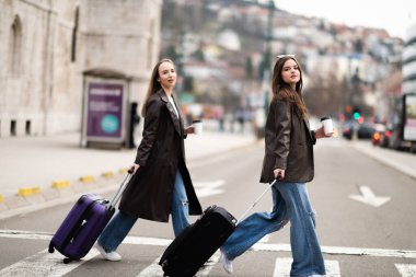 İki şık kadın, ellerinde kahve fincanlarıyla ellerinde bavullarıyla şehir caddesinde yürüyorlar. Kentsel seyahat ve yaşam tarzı konsepti.