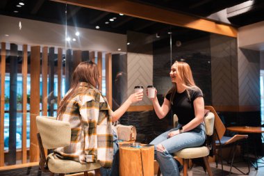 Modern bir kafede oturan iki bayan arkadaş kahve fincanlarıyla kadeh kaldırıyorlar. Sıcak ve dostane atmosfer.