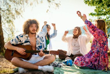 Bir grup arkadaş plajda toplanıp müzik, kahkaha ve birlikteliğin sıcaklığıyla dolu neşeli anları paylaşıyorlar. Gençler sahilde eğleniyor.