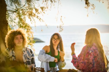 Bir grup arkadaş plajda toplanıp müzik, kahkaha ve birlikteliğin sıcaklığıyla dolu neşeli anları paylaşıyorlar. Gençler sahilde eğleniyor.